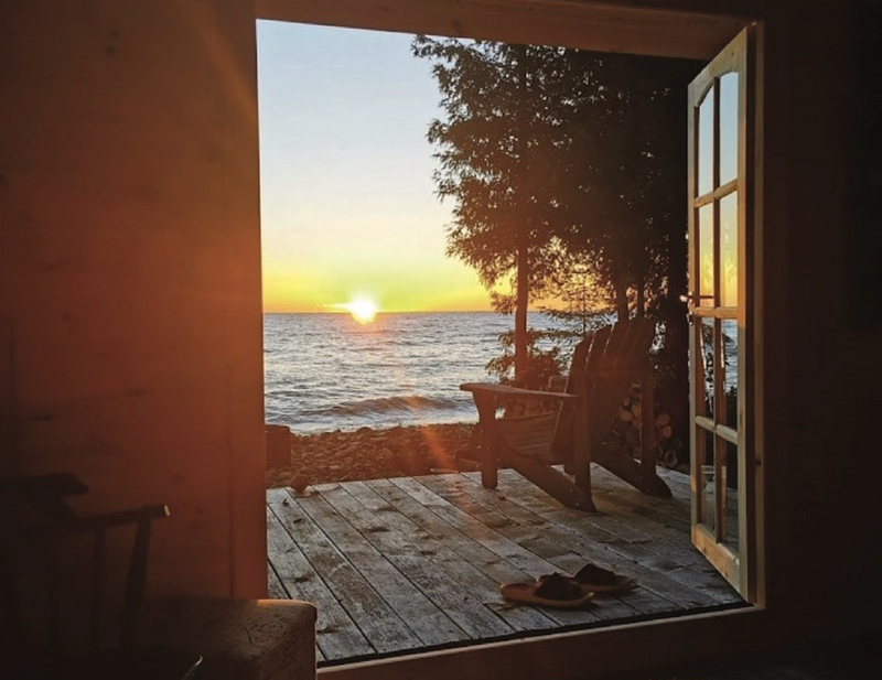 Sunset over water seen through an open window with a wooden bench and chair.